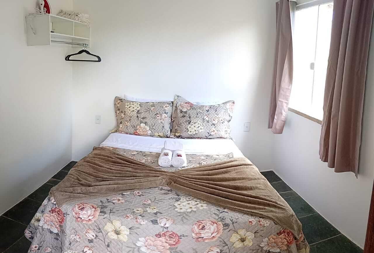 A neatly made bed with floral bedding, a pair of slippers, and a hanging garment rack.
