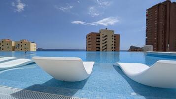 A swimming pool with white lounge chairs, a clear blue sky, and buildings in the background.