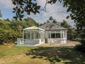 A conservatory with a glass roof, surrounded by a garden and a white building. - Seaview Hafod Y Graig (Talsarnau)