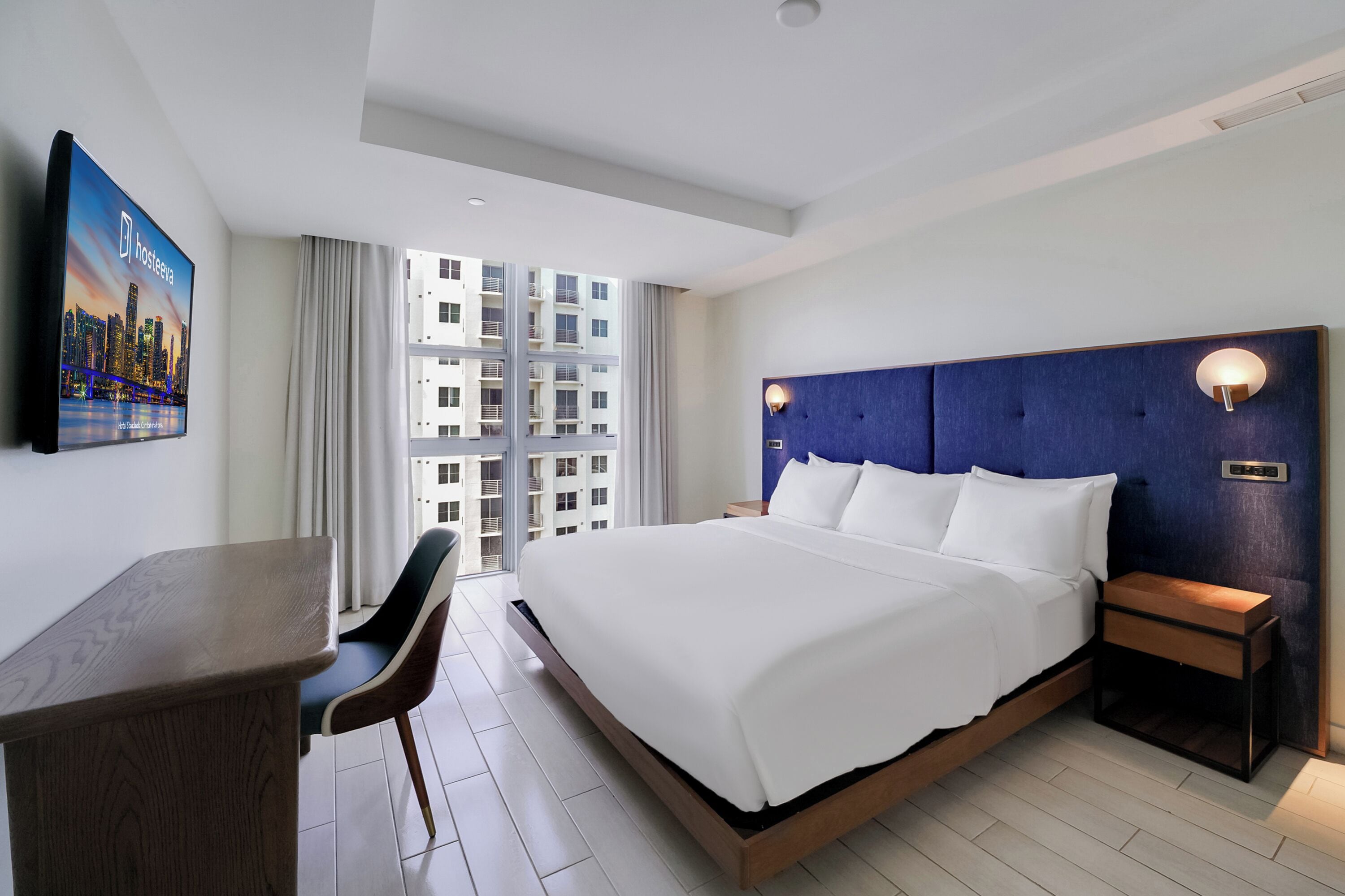 A hotel room with a large bed, a desk, and a chair. There is a TV mounted on the wall and a window with curtains.