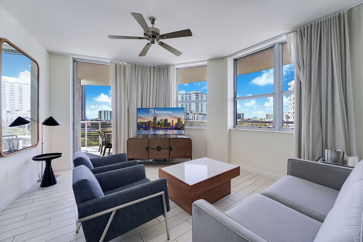 A modern hotel room with a sofa, armchairs, a coffee table, a TV, and large windows offering city views.