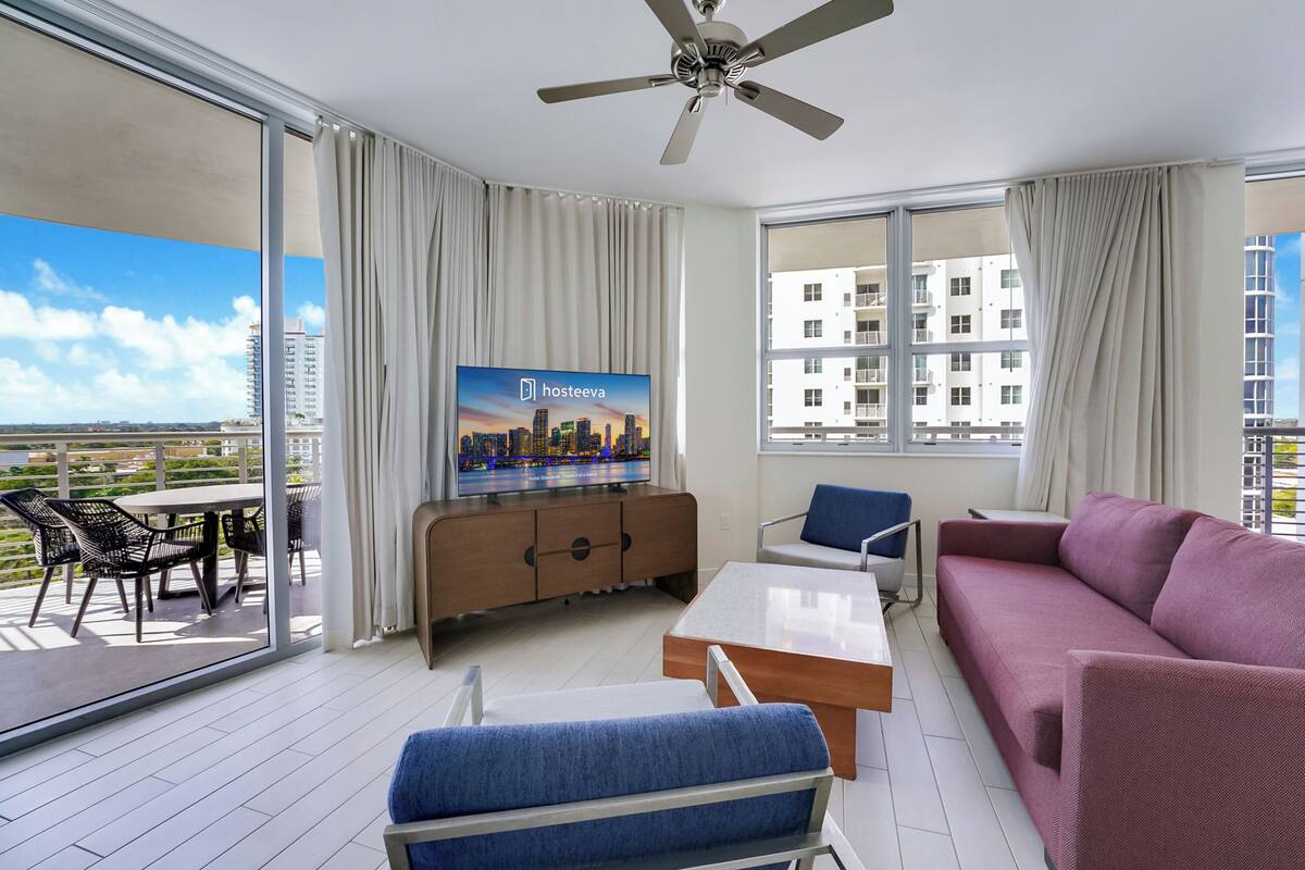 A modern living room with a sofa, chairs, a TV, and a balcony with a city view.