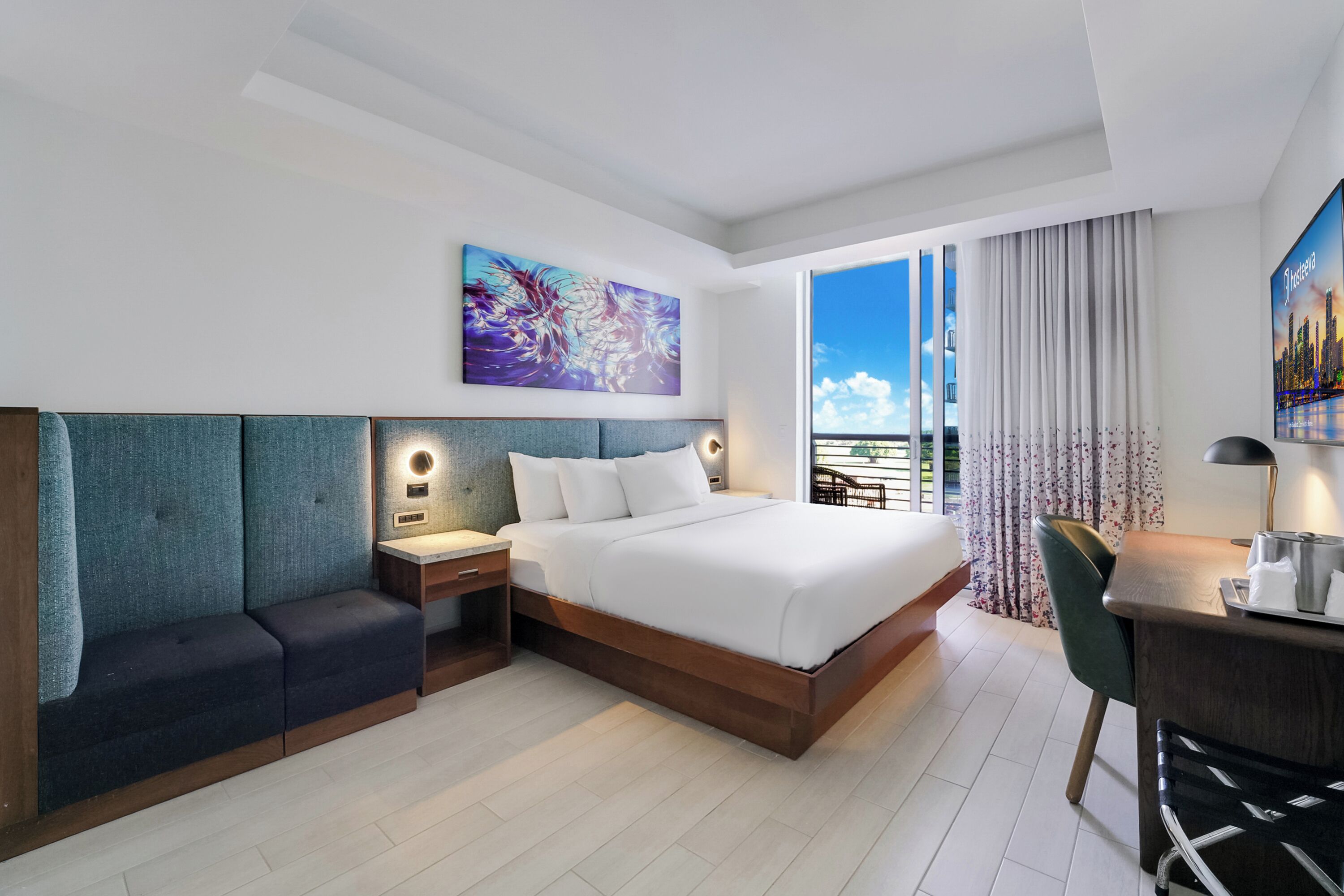A modern hotel room with a large bed, a desk, a chair, a nightstand, and a view of the outdoors.