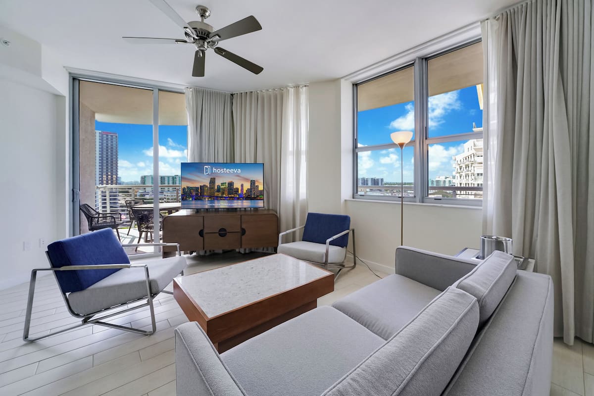 A modern hotel room with a large window, a flat-screen TV, a sofa, a coffee table, and a ceiling fan.
