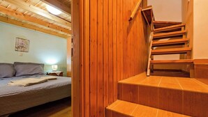 Ein Schlafzimmer mit Bett, Nachttisch und einer Wandlampe. Eine Holztreppe führt zu einer oberen Etage.