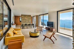 A modern living room with a yellow sofa, a round coffee table, and a television. There is a stone wall and a view of the sea through large windows. - Luxurious Architect-designed Property Pool sea View (Cargese)