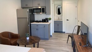 Cocina compacta con armarios grises, refrigerador de acero inoxidable, microondas y cafetera. Cuenta con un pequeño comedor con mesa y sillas, y un espacio de trabajo con computadora y escritorio.