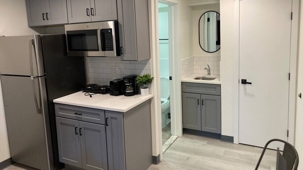 Una cocina compacta con armarios grises, un refrigerador de acero inoxidable, un microondas y una cafetera.