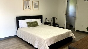 Un letto rifatto con copriletto bianco, cuscino verde e testiera nera in una stanza con pavimento in legno e parete bianca.