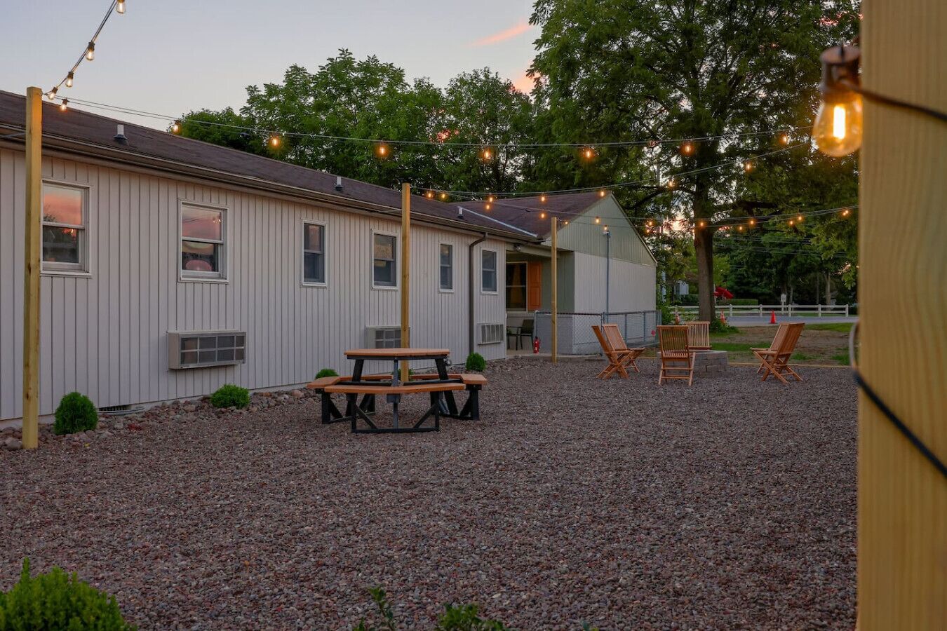 A gravel patio with picnic tables and chairs, a building with a sloped roof, and string lights.
