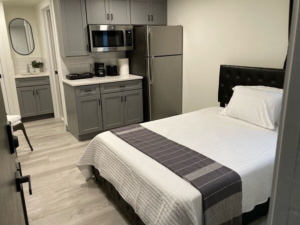 Une chambre d’hôtel de taille réduite, comprenant un lit, une kitchenette équipée d’un four à micro-ondes et d’un réfrigérateur, ainsi qu’une petite salle de bain.