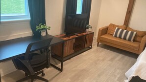 Ein Zimmer mit Schreibtisch, Stuhl, Fernseher und einer Couch mit Kissen.