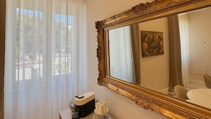 Una habitación de hotel con un espejo grande y ornamentado, una mesita de noche con cafetera y una ventana con cortinas translúcidas.