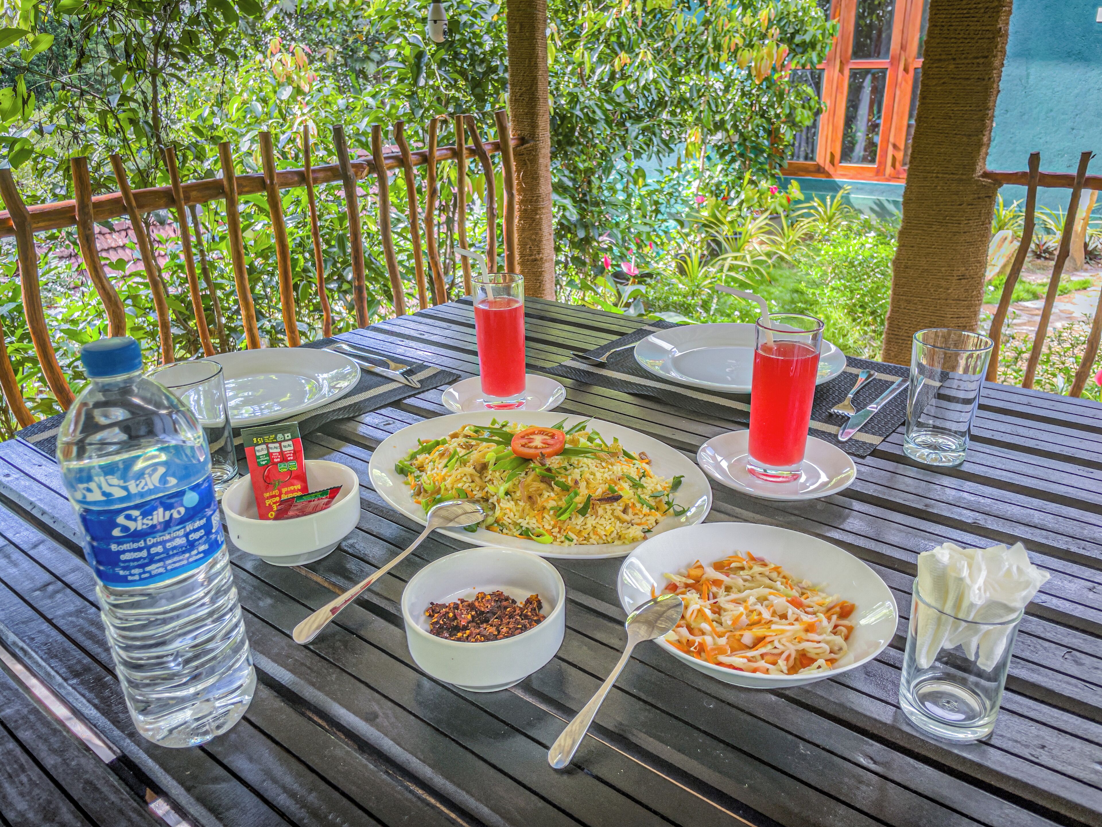 Una mesa dispuesta con platos de comida, una botella de agua y vasos de una bebida roja, rodeada de follaje.