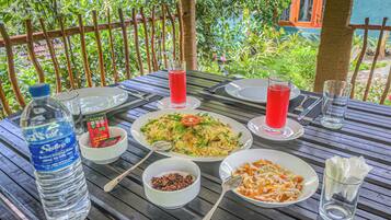 Una mesa dispuesta con platos de comida, una botella de agua y vasos de una bebida roja, rodeada de follaje.