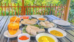 Een tafel gedekt met diverse gerechten, waaronder rijst, curry en een kokosdrank, op een houten tuintafel met uitzicht op weelderig groen.