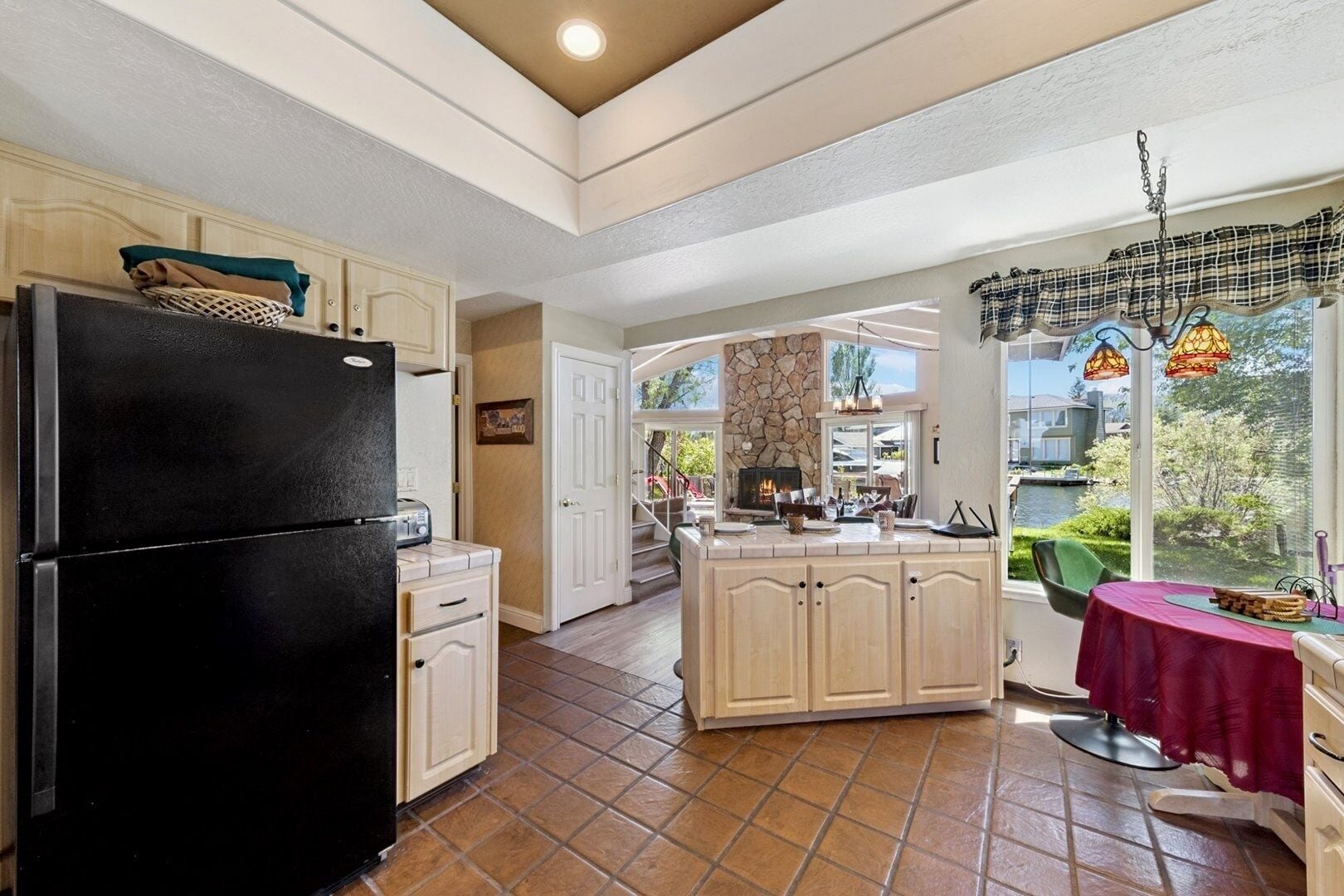 A kitchen with a black refrigerator, a stone fireplace, and a view of a lake.