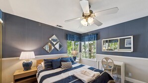 Une chambre à coucher comprenant un lit, une table de chevet, une lampe, un ventilateur de plafond, une fenêtre avec des rideaux et un miroir.