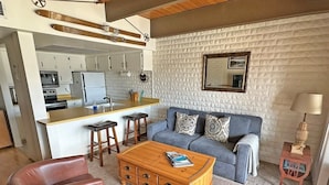 Uma sala de estar moderna com um sofá, mesa de centro e poltronas de couro. Uma cozinha com armários brancos e um balcão é visível ao fundo.