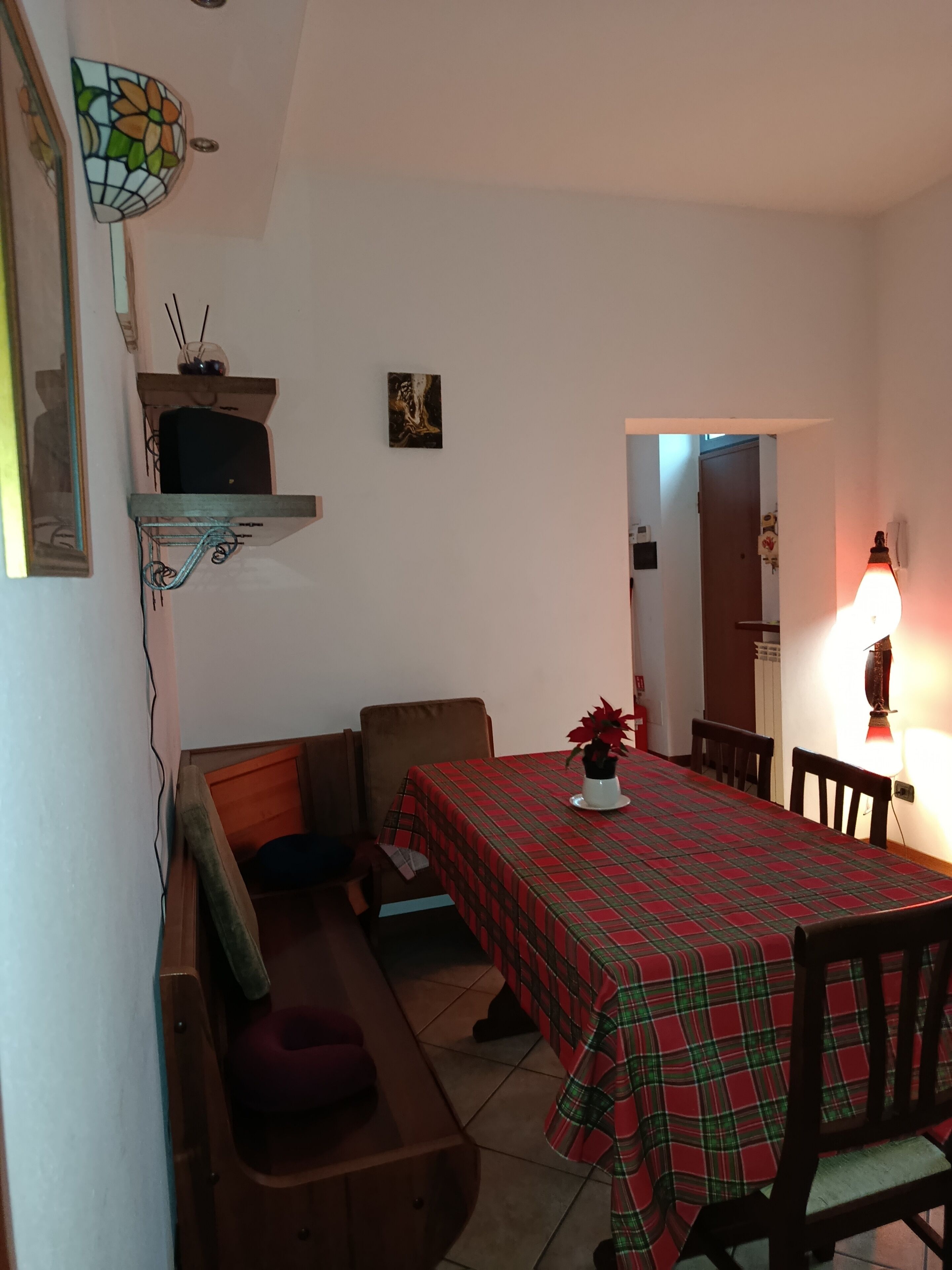 A dining area with a table covered in a plaid tablecloth, chairs, a bench, and a wall-mounted shelf with a television.