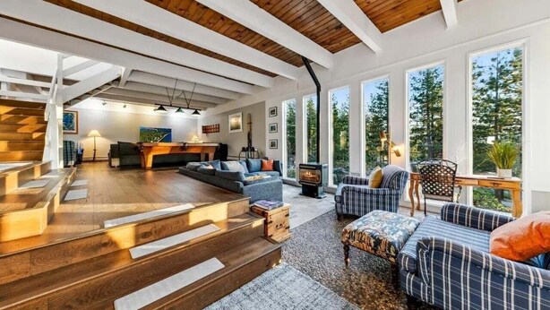 Un amplio salón con escalera de madera, chimenea y grandes ventanales con vistas al exterior.