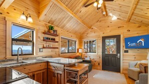 Interior de una cabaña de madera con cocina, comedor y sala de estar. Paredes de madera, una puerta negra y una ventana con vistas.