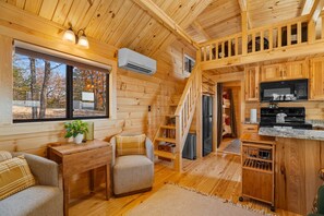 Interior acollidor d'una cabana amb escala de fusta, cuina amb microones i zona de seients amb una planta.