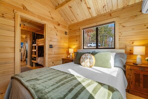Un dormitorio de cabaña de madera con una cama, dos lámparas de noche, un ventanal con vista a árboles y una puerta que da a un altillo.