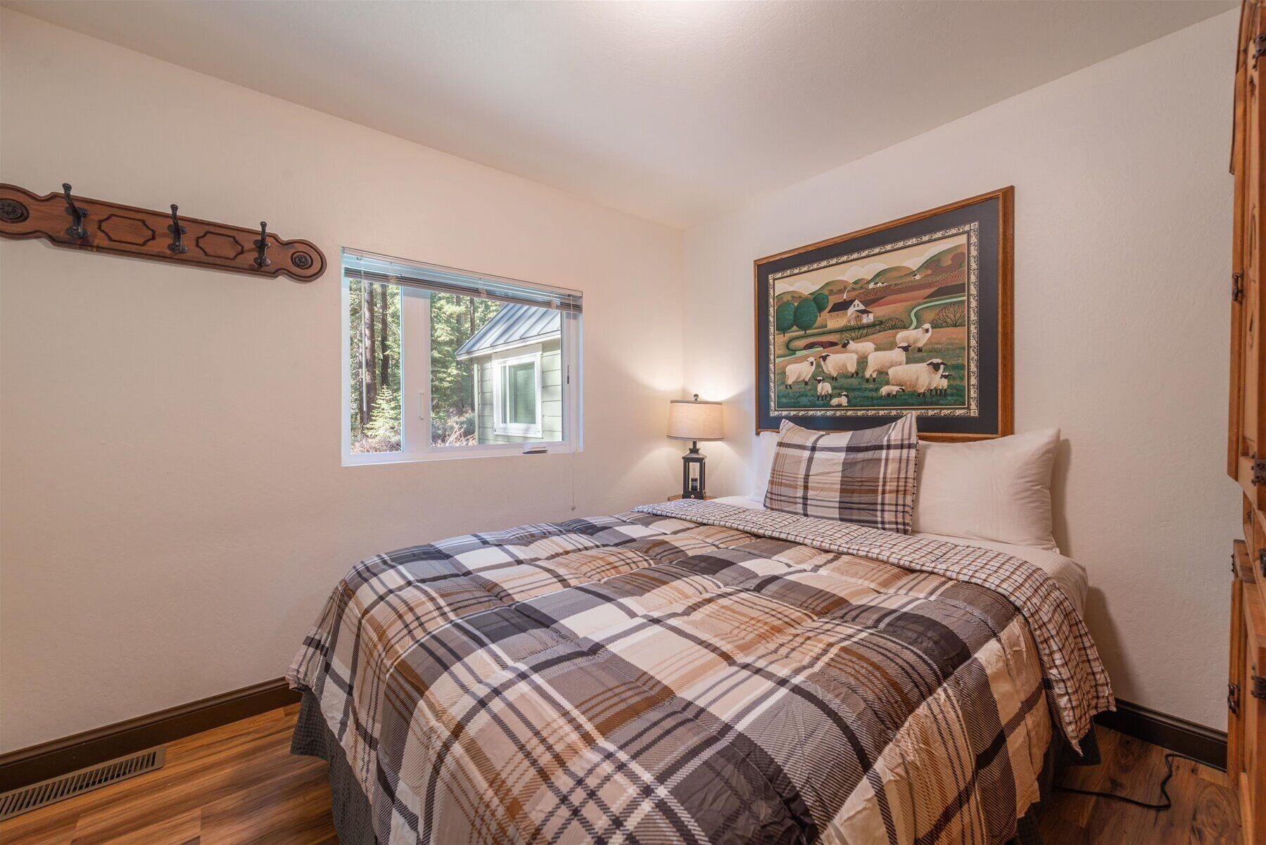 Un dormitorio con una colcha escocesa, una ventana con vistas a una casa y árboles, un cuadro de ovejas y un perchero de madera.