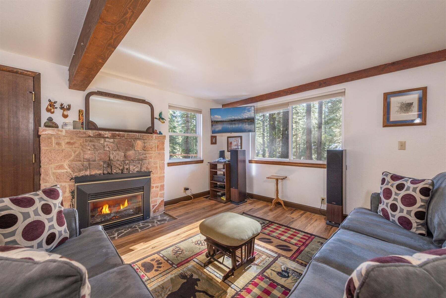 Un salon chaleureux avec une cheminée en pierre, une grande fenêtre donnant sur la forêt et un coin salon confortable agrémenté de coussins à motifs.