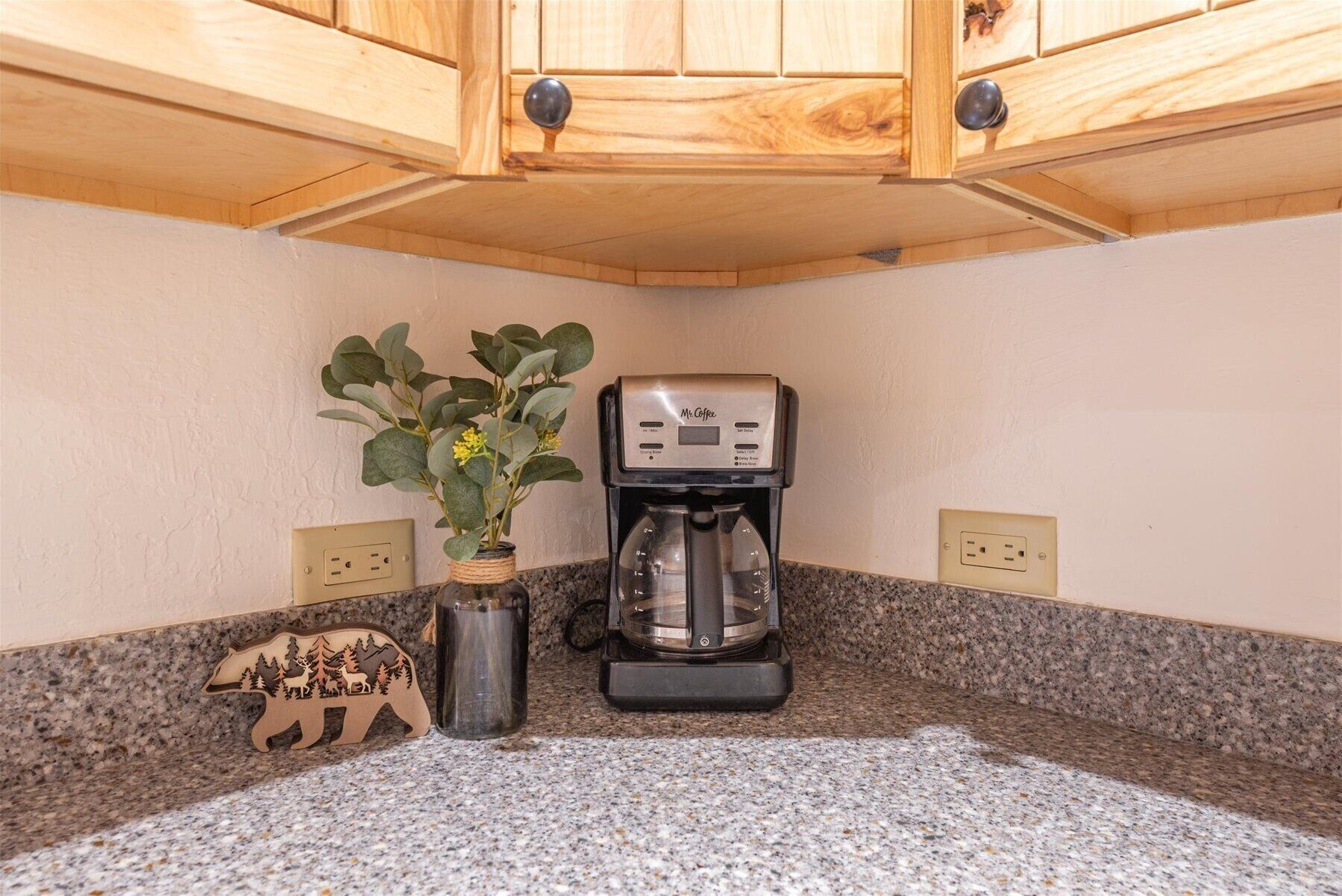 Une cuisine avec des meubles de rangement en bois, une machine à café, un vase rempli de fleurs et une décoration en forme d’ours posée sur le comptoir.