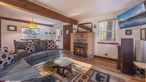 Una sala de estar acogedora con chimenea de piedra, un sofá gris con cojines estampados, suelo de madera y un ventanal con vistas a árboles.