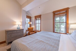 Ein Schlafzimmer mit einem großen Bett, zwei Fenstern mit Jalousien, einem Sessel, einem Schminktisch und zwei Lampen.