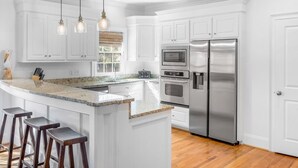 A modern kitchen with white cabinetry, a granite countertop, stainless steel appliances, and a bar stool area. - 6 Bedroom IOP Home w/ Community Pool Just Outside | Steps from Beach and Fun (Isle of Palms)