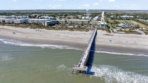 Een luchtbeeld van een kustgebied met een lange pier die het water in reikt, omgeven door gebouwen en een strand.