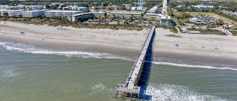 An aerial view of a coastal area with a long pier extending into the water, surrounded by buildings and a beach.