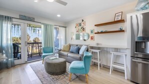 Een moderne woonkamer met een grijze bank, blauwe fauteuils, een rond hocker, een plafondventilator, en een keuken met een koelkast en magnetron.