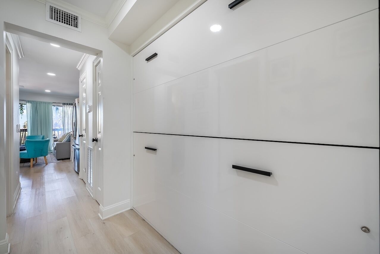 Un couloir avec une armoire blanche, un coin repas avec une chaise bleue et une fenêtre avec des rideaux.