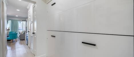Un couloir avec une armoire blanche, un coin repas avec une chaise bleue et une fenêtre avec des rideaux.
