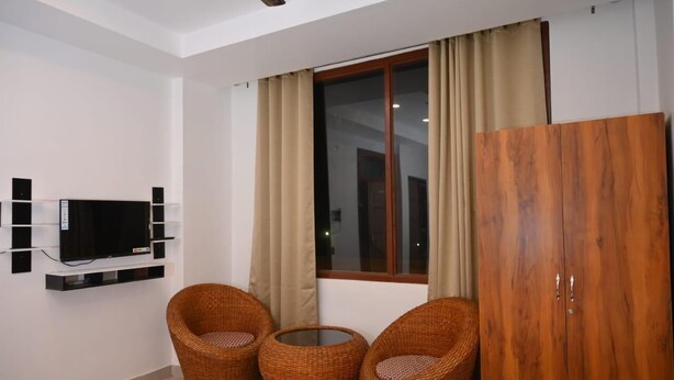 A room with a TV mounted on the wall, a window with curtains, two wicker chairs, a small table, and a wooden wardrobe. - Pitambara Homestay (Faizabad)