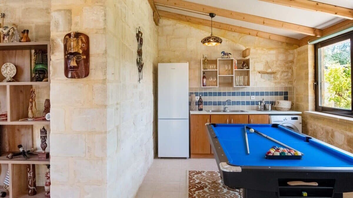 A cozy kitchen with a pool table, stone walls, wooden beams, and a window with a view.