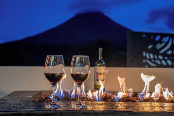 Two glasses of red wine on a wooden table with a bottle and burning wood in the background.