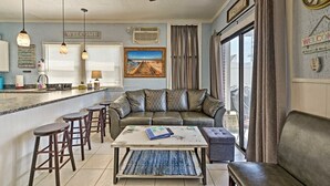 Ein Wohnzimmer mit einer Couch, einem Couchtisch und Barhocker. Es gibt einen Deckenventilator, ein Gemälde und ein Fenster mit Vorhängen.
