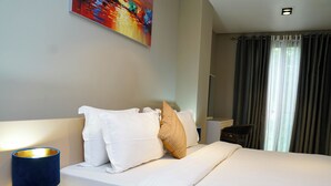 Una camera d'albergo con un letto, biancheria da letto bianca, una lampada da comodino blu, un quadro appeso al muro e una finestra con tende.