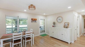 Un vaste espace dédié à la salle à manger, avec une table et des chaises en bois, un buffet blanc agrémenté d’objets décoratifs, et une grande fenêtre donnant sur un paysage verdoyant.