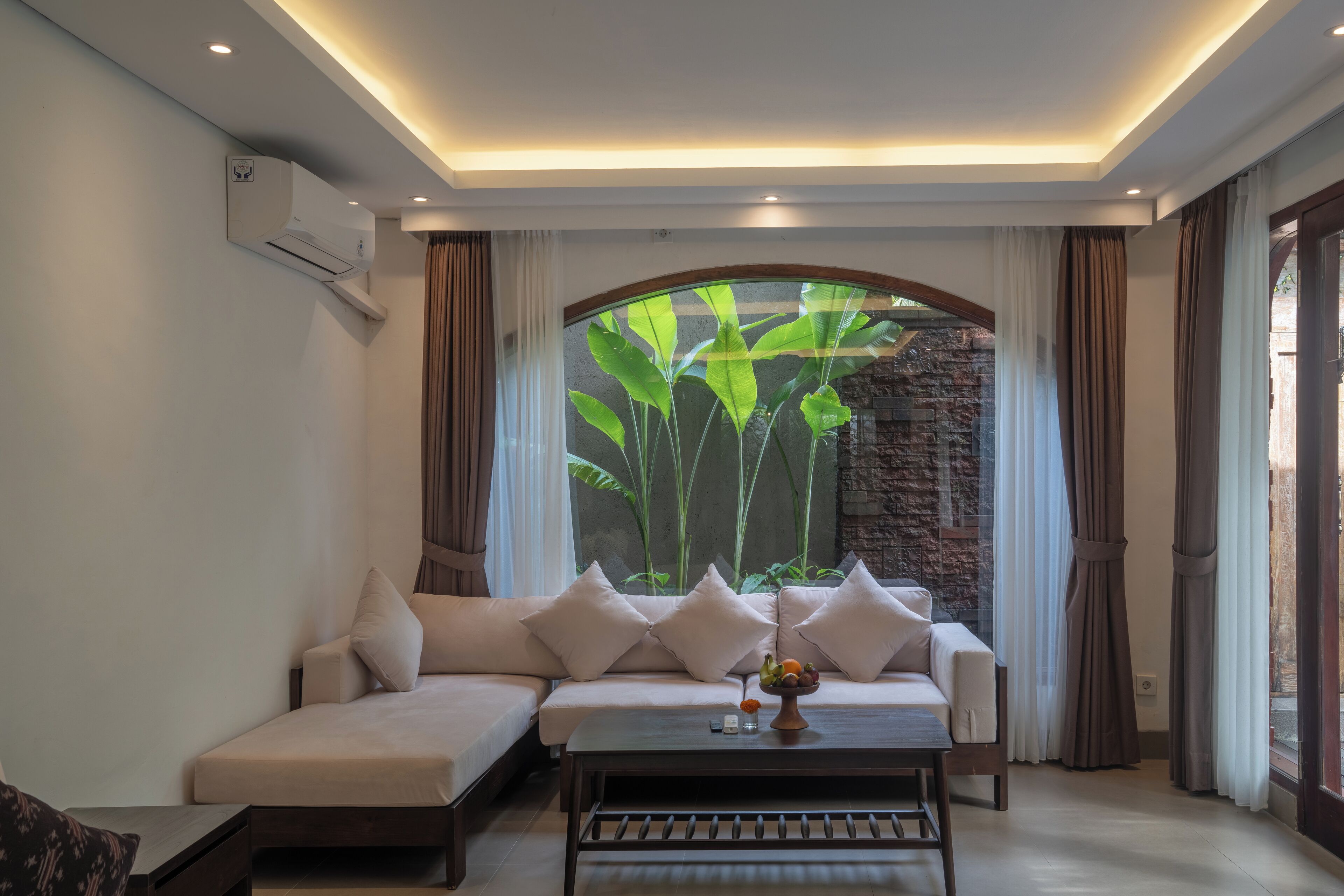 A modern living room with a sofa, a coffee table, and a view of lush greenery through a large window.