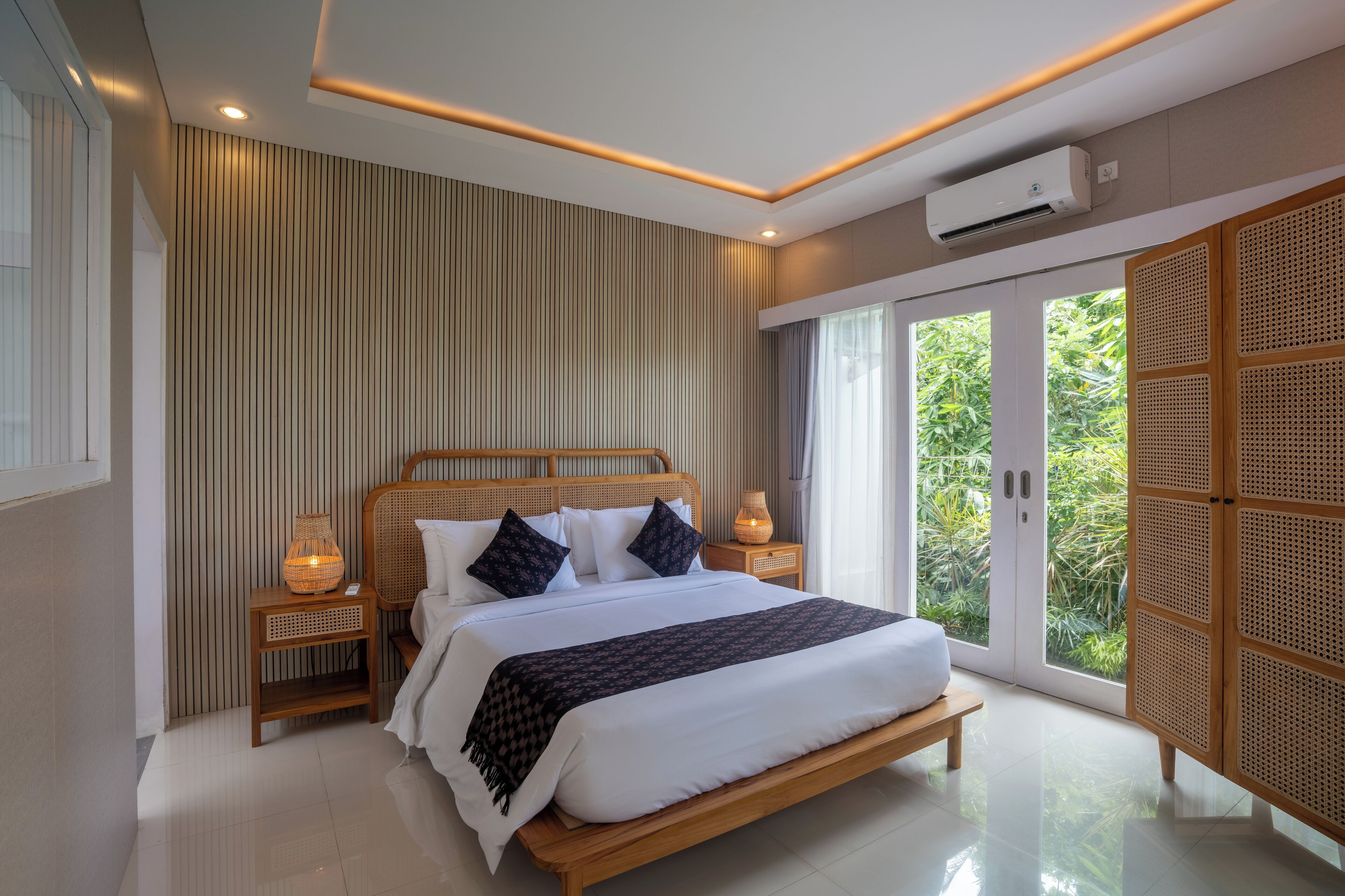 A modern bedroom with a large bed, wooden headboard, and a view through a sliding glass door to a lush garden.