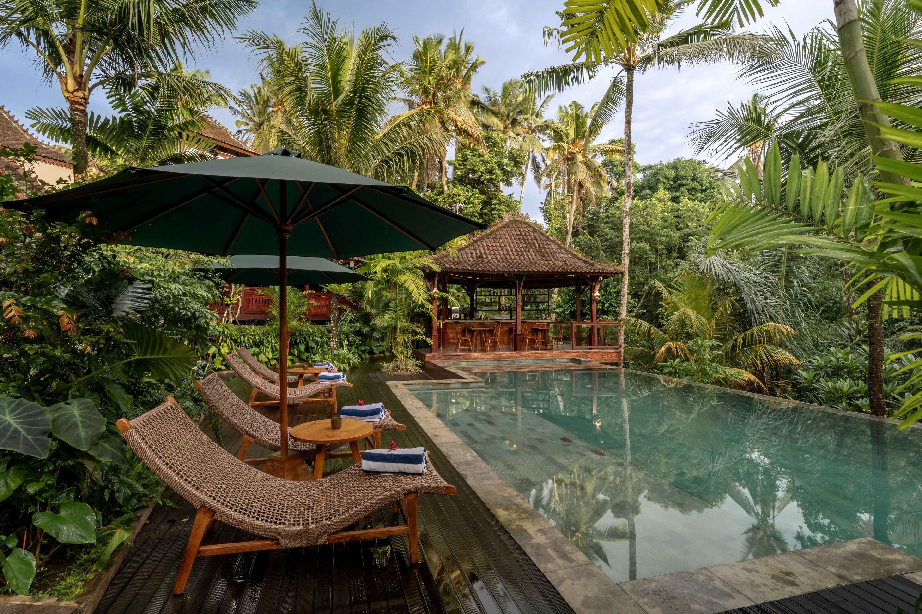 An infinity pool with lounge chairs and umbrellas, surrounded by lush greenery and palm trees.