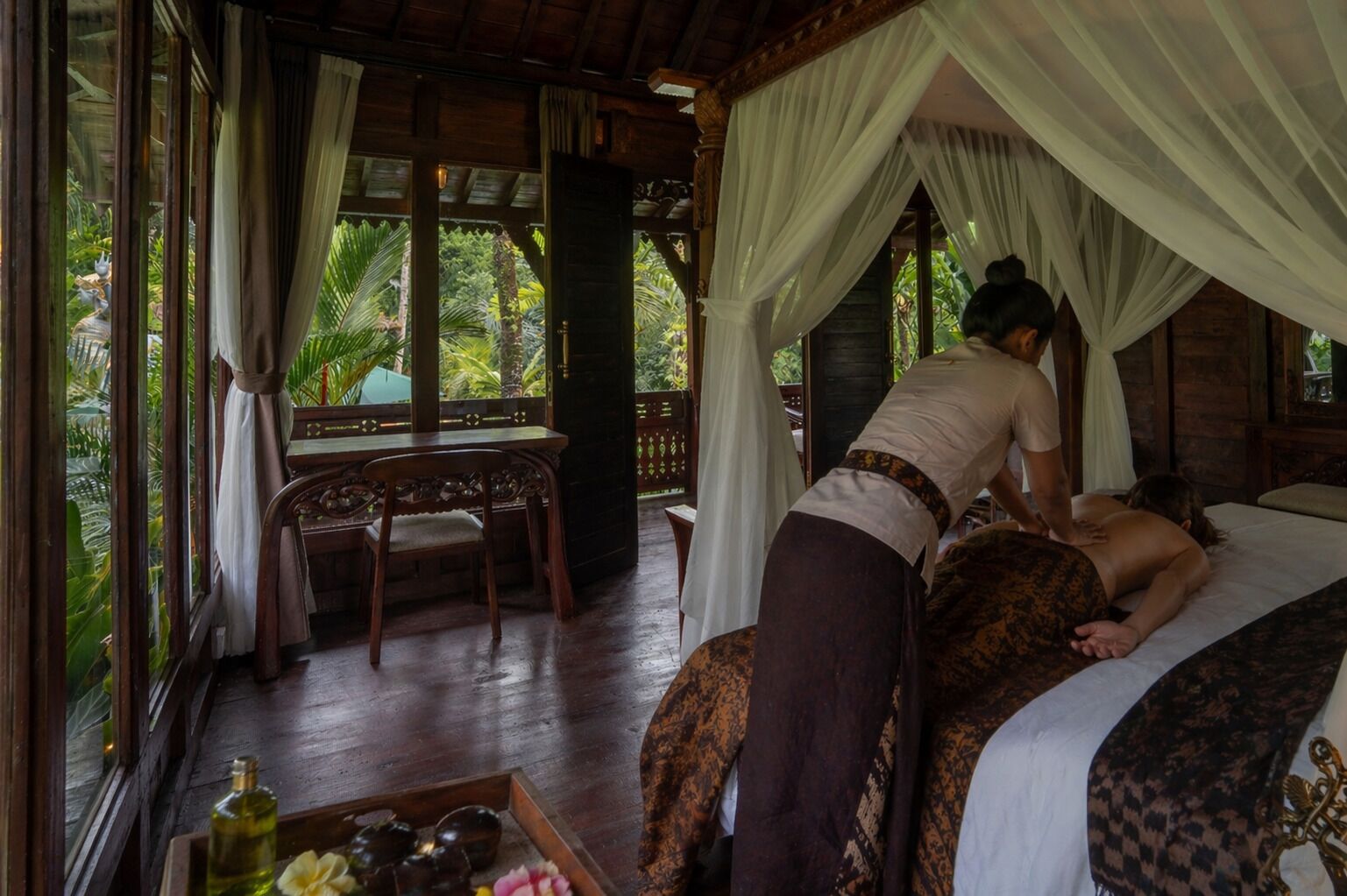 Une pièce comprenant un lit, une table et une chaise. On y voit une personne qui fait un massage à une autre personne allongée sur le lit.