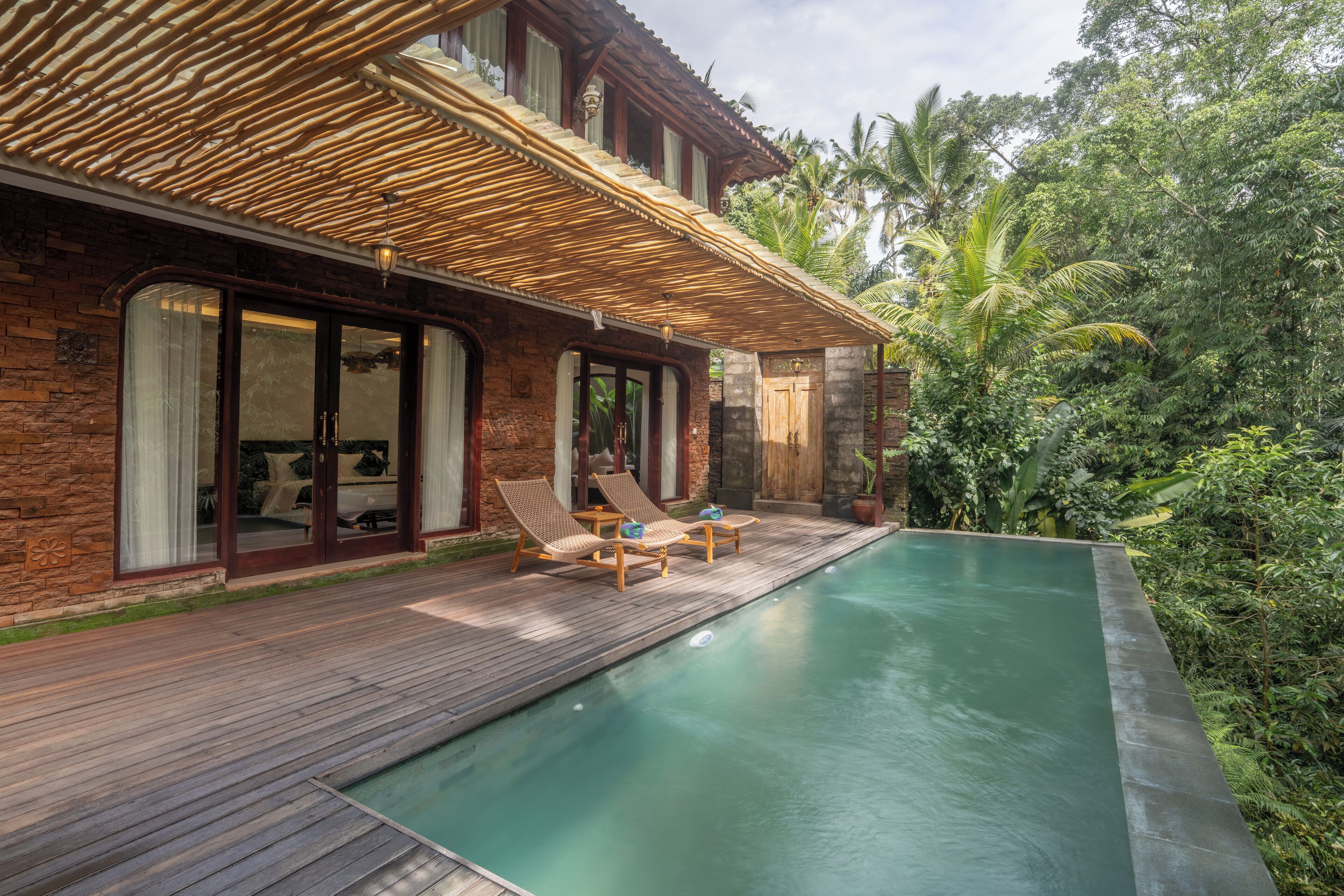 A poolside area with a wooden deck, a pool, and a house with large windows and a wooden roof.